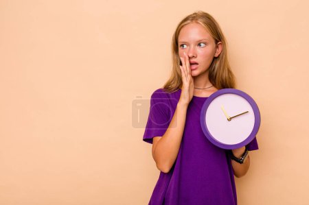 Téléchargez les photos : Petite fille caucasienne tenant une horloge isolée sur fond beige dit une nouvelle secrète de freinage chaud et regardant de côté - en image libre de droit