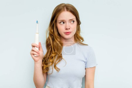 Téléchargez les photos : Jeune femme caucasienne tenant une brosse à dents électrique isolée sur fond bleu confus, se sent douteuse et incertaine. - en image libre de droit