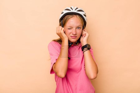 Foto de Niña caucásica usando un casco de bicicleta aislado sobre fondo beige cubriendo las orejas con las manos. - Imagen libre de derechos