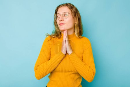 Foto de Joven mujer caucásica aislada sobre fondo azul rezando, mostrando devoción, persona religiosa buscando inspiración divina. - Imagen libre de derechos
