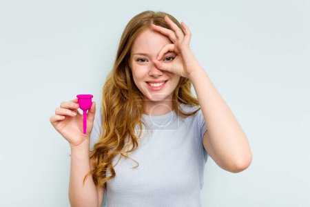 Téléchargez les photos : Jeune femme caucasienne tenant la coupe menstruelle isolée sur fond bleu excité en gardant ok geste sur l'œil. - en image libre de droit