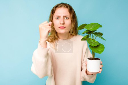Téléchargez les photos : Jeune femme caucasienne tenant une plante isolée sur fond bleu montrant poing à caméra, expression faciale agressive. - en image libre de droit