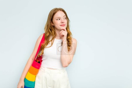 Foto de Joven mujer caucásica sosteniendo bolsa LGTBI aislada sobre fondo azul mirando de lado con expresión dudosa y escéptica. - Imagen libre de derechos