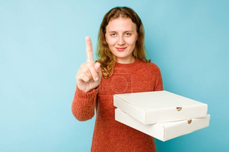 Téléchargez les photos : Jeune femme caucasienne tenant des boîtes de pizzas isolées sur fond bleu montrant numéro un avec doigt. - en image libre de droit