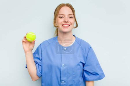 Téléchargez les photos : Jeune kinésithérapeute caucasien tenant une balle de tennis isolée sur fond bleu heureux, souriant et joyeux. - en image libre de droit