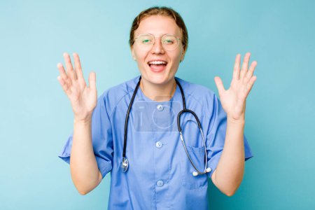 Photo for Young nurse caucasian woman isolated on blue background receiving a pleasant surprise, excited and raising hands. - Royalty Free Image