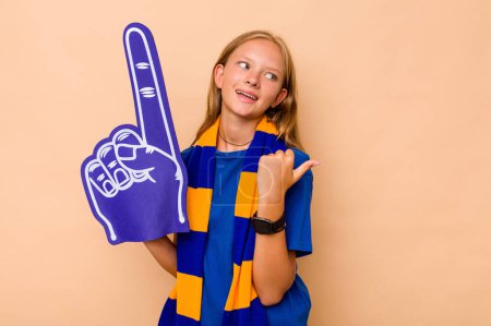 Foto de Poco caucásico deportes fan chica aislado en beige fondo puntos con el dedo pulgar de distancia, riendo y despreocupado. - Imagen libre de derechos