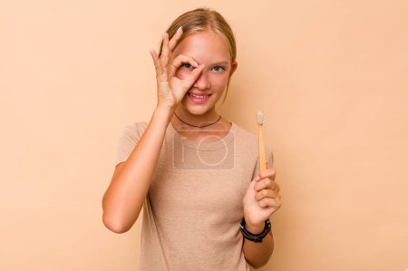 Foto de Caucásico adolescente chica cepillarse los dientes aislado en beige fondo excitado mantener ok gesto en el ojo. - Imagen libre de derechos