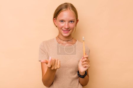 Foto de Caucásico adolescente chica cepillarse los dientes aislado en beige fondo apuntando con el dedo en usted como si invitando a acercarse. - Imagen libre de derechos