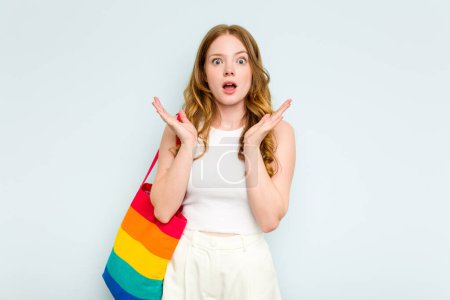 Foto de Joven mujer caucásica sosteniendo bolso LGTBI aislado sobre fondo azul sorprendido y sorprendido. - Imagen libre de derechos