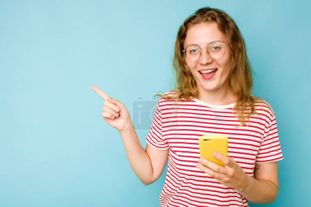 Téléchargez les photos : Jeune femme caucasienne tenant un téléphone portable isolé sur fond bleu souriant et pointant du doigt, montrant quelque chose à l'espace vide. - en image libre de droit