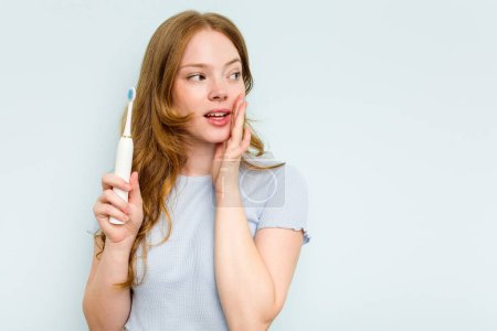 Téléchargez les photos : Jeune femme caucasienne tenant une brosse à dents électrique isolée sur fond bleu dit une nouvelle secrète de freinage chaud et regardant de côté - en image libre de droit