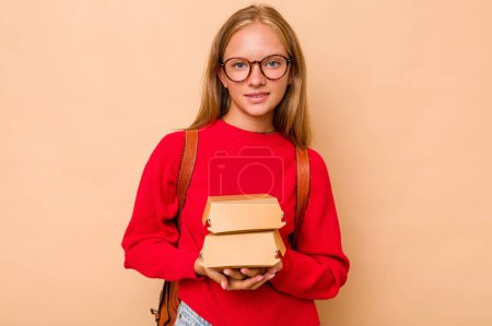 Téléchargez les photos : Petite étudiante caucasienne tenant des hamburgers isolés sur fond beige heureuse, souriante et joyeuse. - en image libre de droit