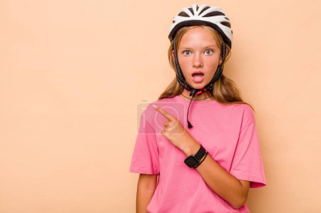Téléchargez les photos : Petite fille caucasienne portant un casque de vélo isolé sur fond beige pointant vers le côté - en image libre de droit