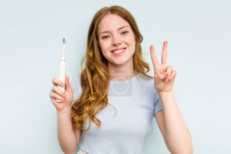 Téléchargez les photos : Jeune femme caucasienne tenant une brosse à dents électrique isolée sur fond bleu montrant le numéro deux avec les doigts. - en image libre de droit