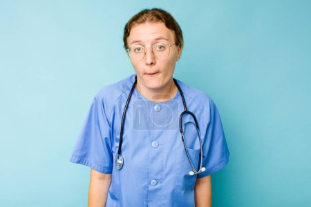 Téléchargez les photos : Jeune infirmière caucasienne isolée sur fond bleu haussant les épaules et ouvrant les yeux confus. - en image libre de droit