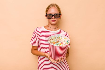 Photo for Little caucasian girl eating popcorn isolated on beige background shrugs shoulders and open eyes confused. - Royalty Free Image