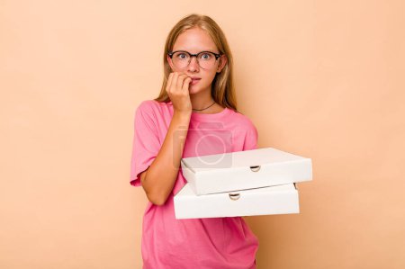 Téléchargez les photos : Petite fille caucasienne tenant une pizza isolée sur fond beige mordant les ongles, nerveuse et très anxieuse. - en image libre de droit