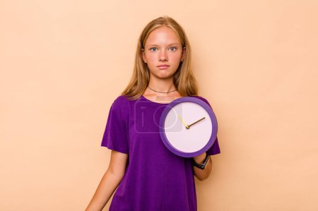 Foto de Niña caucásica sosteniendo un reloj aislado sobre fondo beige encoge hombros y los ojos abiertos confundido. - Imagen libre de derechos
