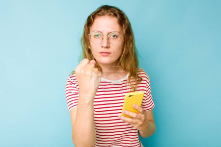 Téléchargez les photos : Jeune femme caucasienne tenant téléphone portable isolé sur fond bleu montrant poing à caméra, expression faciale agressive. - en image libre de droit
