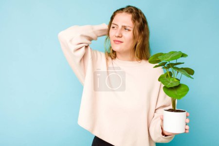 Téléchargez les photos : Jeune femme caucasienne tenant une plante isolée sur fond bleu touchant l'arrière de la tête, pensant et faisant un choix. - en image libre de droit