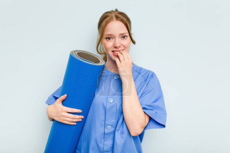 Téléchargez les photos : Jeune kinésithérapeute caucasien tenant un tapis isolé sur fond bleu les ongles mordants, nerveux et très anxieux. - en image libre de droit