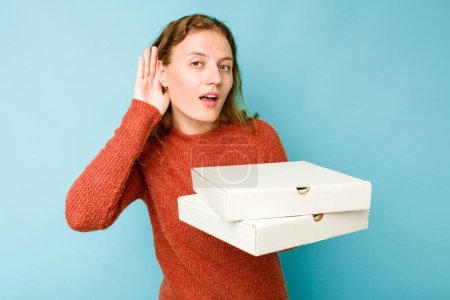 Photo for Young caucasian woman holding pizzas boxes isolated on blue background trying to listening a gossip. - Royalty Free Image