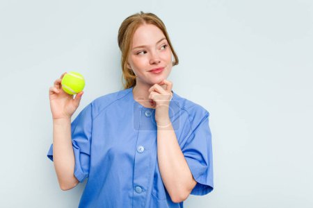Foto de Joven fisioterapeuta caucásica sosteniendo una pelota de tenis aislada sobre fondo azul mirando de lado con expresión dudosa y escéptica. - Imagen libre de derechos