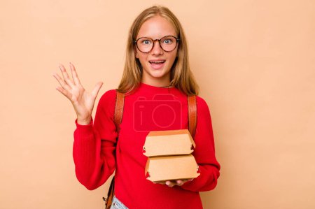 Photo for Little caucasian student girl holding burgers isolated on beige background receiving a pleasant surprise, excited and raising hands. - Royalty Free Image