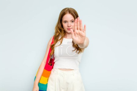 Foto de Mujer joven caucásica sosteniendo bolsa LGTBI aislada sobre fondo azul de pie con la mano extendida mostrando señal de stop, lo que le impide. - Imagen libre de derechos