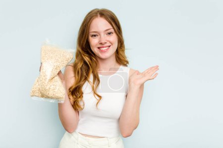 Photo for Young caucasian woman holding oatmeal isolated on blue background showing a copy space on a palm and holding another hand on waist. - Royalty Free Image