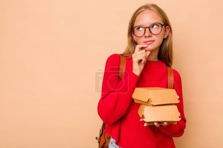 Photo for Little caucasian student girl holding burgers isolated on beige background looking sideways with doubtful and skeptical expression. - Royalty Free Image