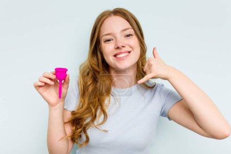 Foto de Joven mujer caucásica sosteniendo copa menstrual aislada sobre fondo azul mostrando un gesto de llamada telefónica móvil con los dedos. - Imagen libre de derechos