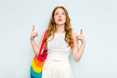 Foto de Joven mujer caucásica sosteniendo bolsa LGTBI aislada sobre fondo azul apuntando al revés con la boca abierta. - Imagen libre de derechos