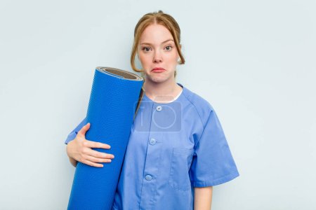 Foto de Joven fisioterapeuta caucásica sosteniendo una esterilla aislada sobre fondo azul encoge hombros y ojos abiertos confundidos. - Imagen libre de derechos