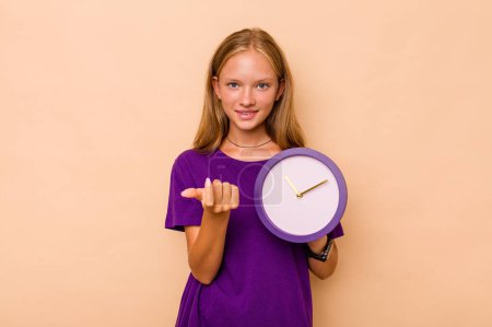 Photo for Little caucasian girl holding a clock isolated on beige background pointing with finger at you as if inviting come closer. - Royalty Free Image