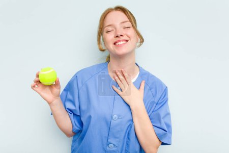 Téléchargez les photos : Jeune kinésithérapeute caucasien tenant une balle de tennis isolée sur fond bleu rit bruyamment en gardant la main sur la poitrine. - en image libre de droit