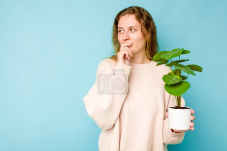 Photo for Young caucasian woman holding a plant isolated on blue background looking sideways with doubtful and skeptical expression. - Royalty Free Image