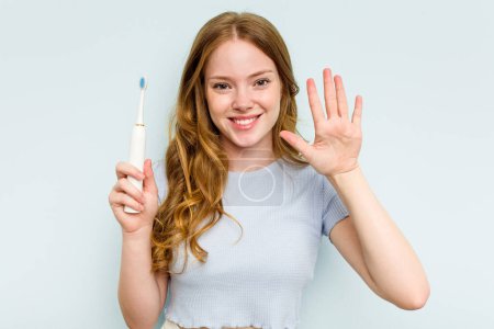 Téléchargez les photos : Jeune femme caucasienne tenant une brosse à dents électrique isolée sur fond bleu souriant gai montrant numéro cinq avec les doigts. - en image libre de droit