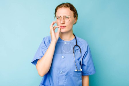 Téléchargez les photos : Jeune infirmière caucasienne isolée sur fond bleu avec les doigts sur les lèvres gardant un secret. - en image libre de droit