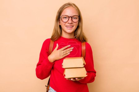 Téléchargez les photos : Petite étudiante caucasienne tenant des hamburgers isolés sur fond beige rit bruyamment en gardant la main sur la poitrine. - en image libre de droit