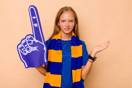 Téléchargez les photos : Petite fille fan de sport caucasienne isolée sur fond beige montrant un espace de copie sur une paume et tenant une autre main sur la taille. - en image libre de droit