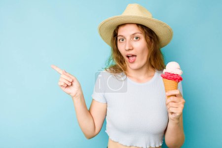 Foto de Joven mujer caucásica sosteniendo un helado aisló un fondo azul apuntando hacia un lado - Imagen libre de derechos