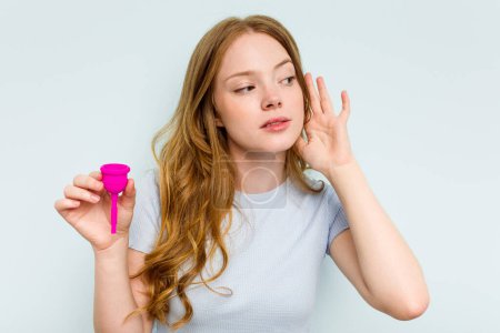 Téléchargez les photos : Jeune femme caucasienne tenant la coupe menstruelle isolée sur fond bleu essayant d'écouter un potins. - en image libre de droit