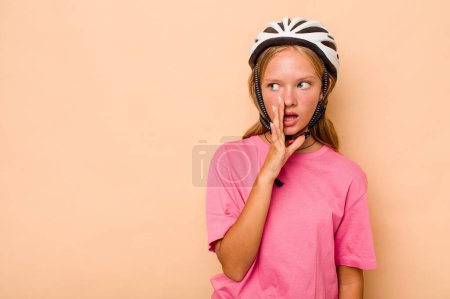 Foto de Pequeña chica caucásica con un casco de bicicleta aislado sobre fondo beige está diciendo una noticia secreta de frenado caliente y mirando a un lado - Imagen libre de derechos