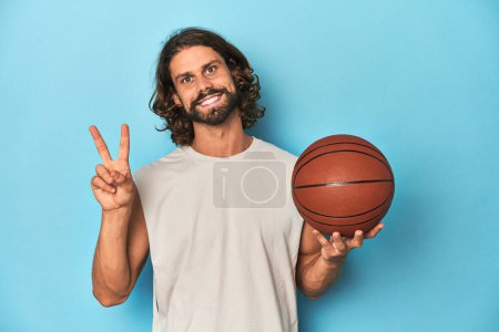 Foto de Hombre barbudo con baloncesto en estudio azul alegre y despreocupado mostrando un símbolo de paz con los dedos. - Imagen libre de derechos
