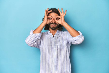 Photo for Man with beard in blue striped shirt, blue studio showing okay sign over eyes - Royalty Free Image