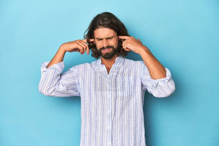 Photo for Man with beard in blue striped shirt, blue studio covering ears with fingers, stressed and desperate by a loudly ambient. - Royalty Free Image