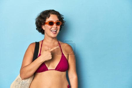 Photo for Young woman in bikini with beach bag smiling and pointing aside, showing something at blank space. - Royalty Free Image