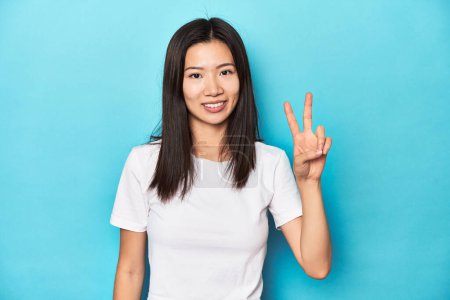 Téléchargez les photos : Jeune femme asiatique en t-shirt blanc, plan studio, joyeuse et insouciante montrant un symbole de paix avec les doigts. - en image libre de droit